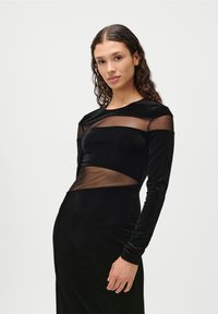 Garā melna kleita no samta, ar caurspīdīgiem sieta paneļiem plecos un jostasvietā. Pieguļošs augšdaļa ar garām piedurknēm un gludu tekstūru.