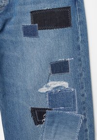 Blauwe spijkerbroek met meerdere vierkante patches in verschillende tinten en texturen, waaronder donker indigo en rafelige randen, wat zorgt voor een verweerde uitstraling.