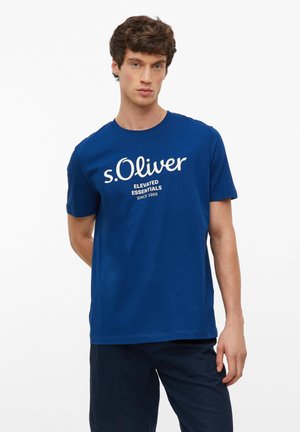 Jonge man met krullend haar die een blauw s.Oliver t-shirt met witte tekst draagt, staand tegen een effen witte achtergrond.