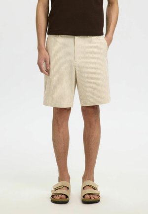 Shorts rayés beige et blanc avec une coupe ample, fabriqués en tissu léger. Associés à une chemise marron et des sandales slides beiges.