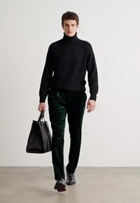 Black turtleneck sweater, green velvet pants, carrying a structured black bag, with dark shoes, walking against a light background.