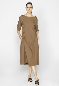 Brązowa sukienka do kolan z rękawami do trzech czwarte, kieszeniami i dekoltem łódkowym; gładki materiał; modelka ma na sobie czarne sandały.
