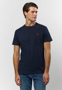 Námořnická modrá bavlněná trička s kulatým výstřihem, krátkými rukávy a malým červeným vyšitým logem na hrudi. Měkká textura, regular fit.