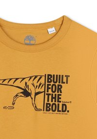 Geel shirt met een zwarte dinosaurusafbeelding en de tekst "Bouwt voor de Dapperen. Sinds 1973, maar misschien eerder," Timberland-logo zichtbaar.