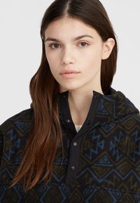 Pull en polaire avec des motifs géométriques en vert foncé et bleu. Il dispose d'une patte de boutonnage noire avec des snaps et d'une poche avant. Logo de la marque visible.