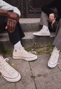 Beige canvas high-top sneakers with white rubber soles and red accents. Features metal eyelets and a circular logo on the side.
