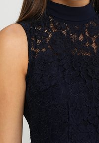 Ärmös mörkblå topp med blommig spetsöverlägg och hög hals, bärs av en person med rakt brunt hår.