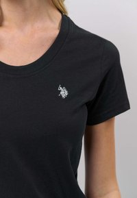 Črna bombažna majica z okroglim izrezom, z majhnim belim vezenim logotipom na levi strani prsi. Kratki rokavi, gladka tekstura.