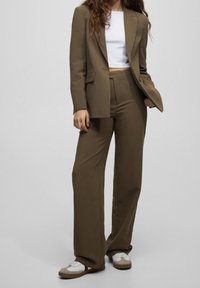 Costume vert olive ajusté avec une veste à coupe décontractée et un pantalon à jambes larges, accompagné d'un top blanc court et de baskets blanches avec des accents marron.