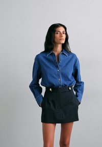 Denim skjorte med knapper i blått parret med et høydejustert svart skjørt med gullfarget detaljer. Glatt tekstur, skreddersydd passform.