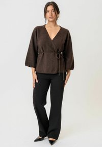 Brauner Wickelsweater mit tiefem V-Ausschnitt, drei-viertel langen Ärmeln und einem Taillengürtel, kombiniert mit schwarzen weiten Hosen und spitzen Schuhen.