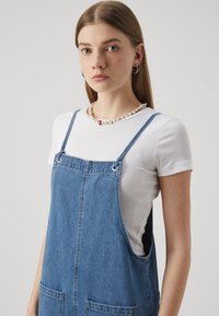 Denim overall obleka z dvema sprednjima žepoma, nastavljivima naramnicama in svetlo pranjeno. Oblečena preko prilegajoče bele rebraste majice.