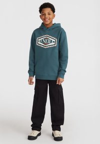 Felpa color teal con logo decorativo, abbinata a pantaloni cargo neri. Presenta un cappuccio con coulisse, polsini a costine e sneakers bianco sporco.