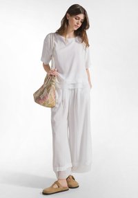 Vrouw in losse witte broek en overhemd met een schoudertas met patroon, draagend beige instappers, staand tegen een witte achtergrond.