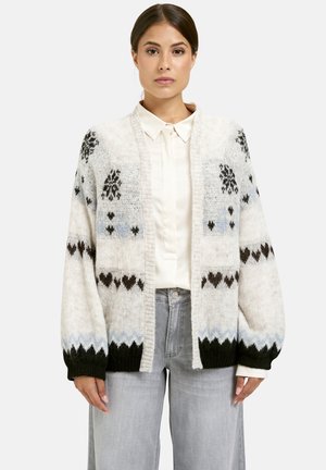 Eine Frau trägt eine cremefarbene Strickjacke mit schwarzen und blauen geometrischen Mustern über einem weißen Hemd mit Kragen und hellgrauen Hosen.