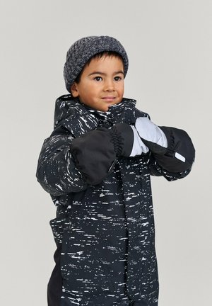 Veste d'hiver noire avec des motifs abstraits blancs, dotée d'une capuche et de poignets élastiques. Un bonnet tricoté gris chaud et des moufles assorties sont portés.