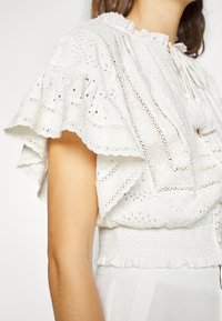 Weißes besticktes Blusenshirt mit kurzen Rüschensäumen, filigranen Lochmustern und schmaler Taille. Mit einer Bindung am Ausschnitt.