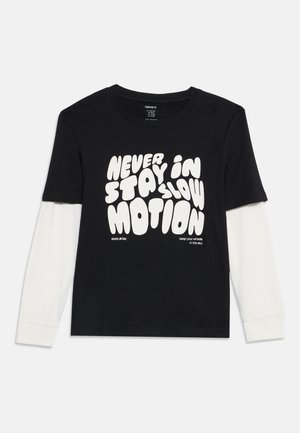 T-shirt à manches longues noir et blanc avec un texte blanc en gras indiquant "Never stay in slow motion" et un texte plus petit à propos du skateboard et des roues.