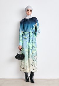 Langes Blumenkleid in Farbverlaufsschattierungen von Blau und Grün, mit gerafftem Ausschnitt und tailliertem Bund. Kombiniert mit einer schwarzen Handtasche und spitzen Stiefeln.
