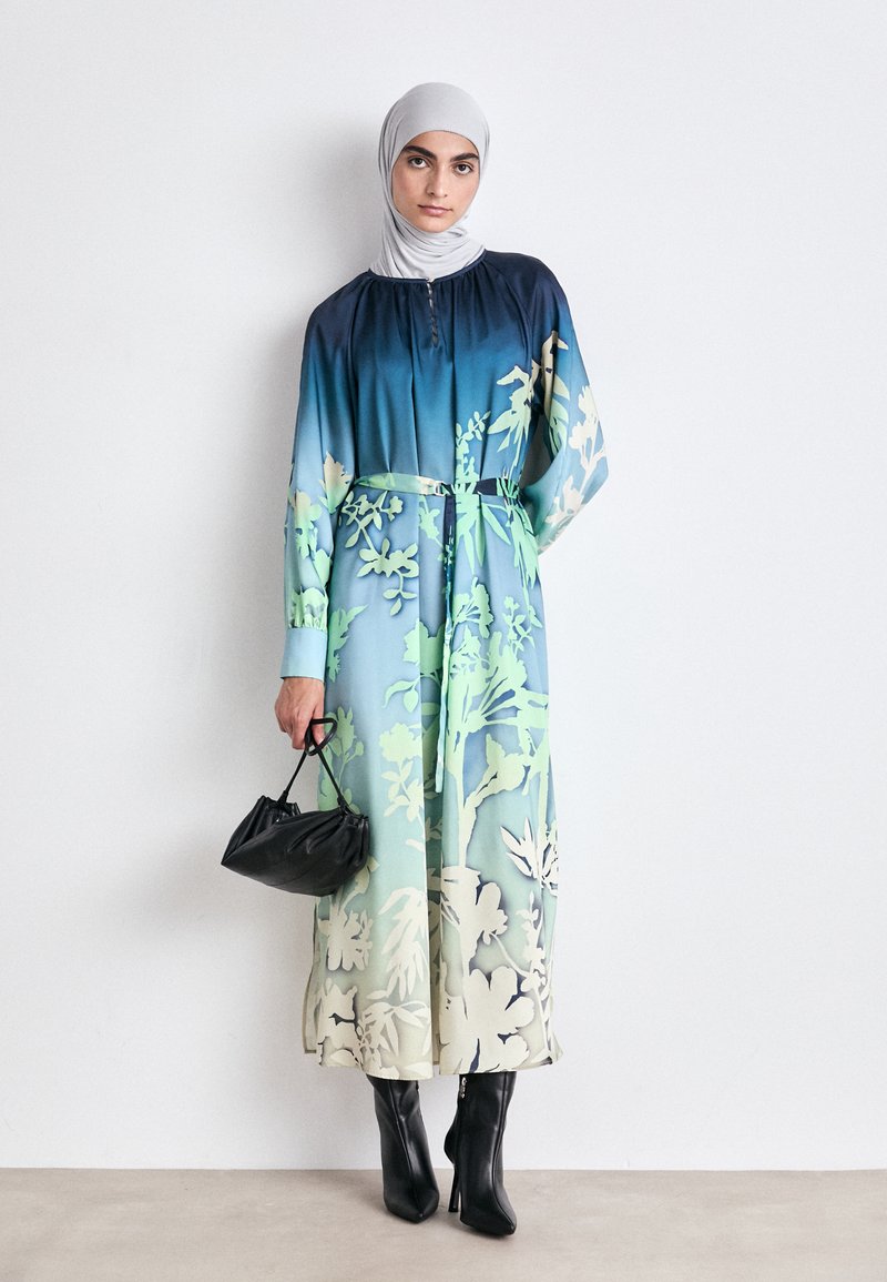 Langes Blumenkleid in Farbverlaufsschattierungen von Blau und Grün, mit gerafftem Ausschnitt und tailliertem Bund. Kombiniert mit einer schwarzen Handtasche und spitzen Stiefeln.
