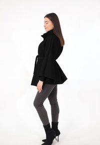 Schwarzer Mantel mit hohem Kragen und weitem Ärmel. Mit einem Taillengürtel, glatter Textur und reicht bis zur Hüfte, kombiniert mit enganliegenden grauen Hosen.