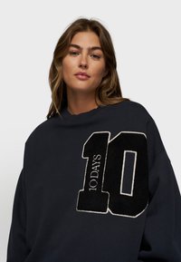 Felpa blu marina con un grande numero "10" in rilievo di colore nero, bordato di bianco, e la scritta "10 GIORNI". Collo a giro e vestibilità comoda.