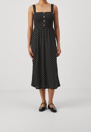 Vrouw draagt een zwarte mouwloze midi-jurk met witte stippen en knopen op het lijfje, gecombineerd met zwarte Mary Jane schoenen.