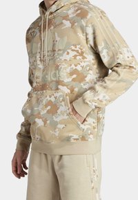 Sudadera beige con patrón de camuflaje, con bolsillo integrado, capucha con cordones y logo de Adidas en tono. Fabricada en tejido suave.