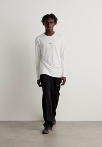 T-shirt branca de manga longa com uma marcação subtil, combinada com calças largas pretas e sapatos escuros, em frente a um fundo claro e liso.