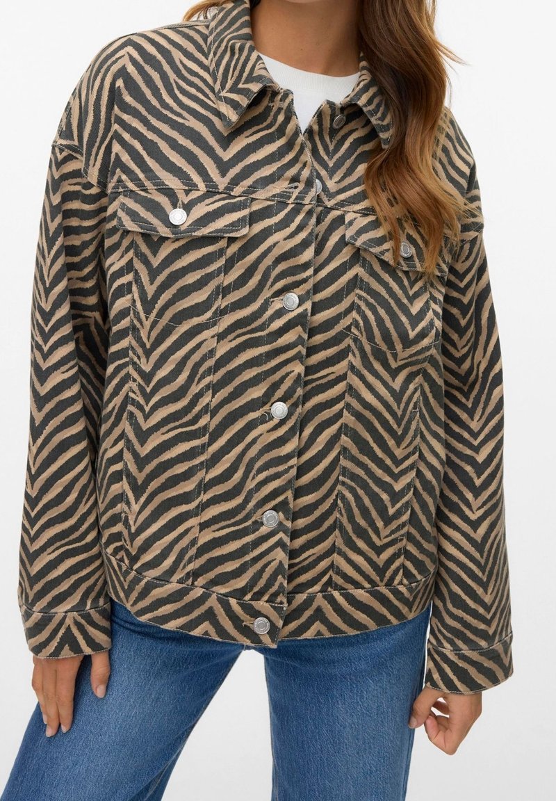 Femme portant une veste boutonnée marron et noire à rayures zèbre avec des poches à rabat sur la poitrine et un jean en denim bleu.