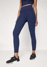 Sötétkék, magas derekú leggings, rugalmas anyagból készült, lapos varratokkal és tapered (szűkülő) fazonnal. Fekete sportcipővel párosítva.