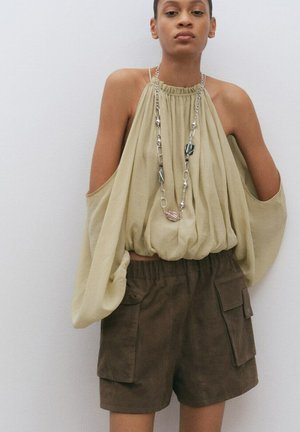 Jeune personne portant un haut beige fluide à épaules dénudées, un long collier, et un short cargo marron, debout devant un mur clair uni.