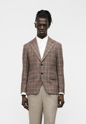 Man met dreadlocks, gekleed in een bruin ruitjesblazer over een wit overhemd en beige broek, staand tegen een effen lichte achtergrond.