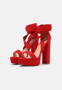Chaussures à talons hauts en daim rouge avec des brides croisées à la cheville et un talon compensé épais, présentant un design à bout ouvert.