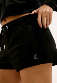 Gros plan sur une personne portant un short noir avec une poche zippée et un petit logo en strass sur l'ourlet, la main posée sur la taille.