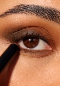 Close-up of a brown eye with eyeliner being applied. The iris is rich brown, surrounded by light eyelashes and soft, warm skin tones.