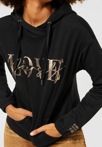 Svart huvtröja med en cowl-neck, prydd med metallisk guldtext "ÄLSKA dig själv" och ett citat på ärmen. Tyget hänger löst och har en mjuk konsistens.