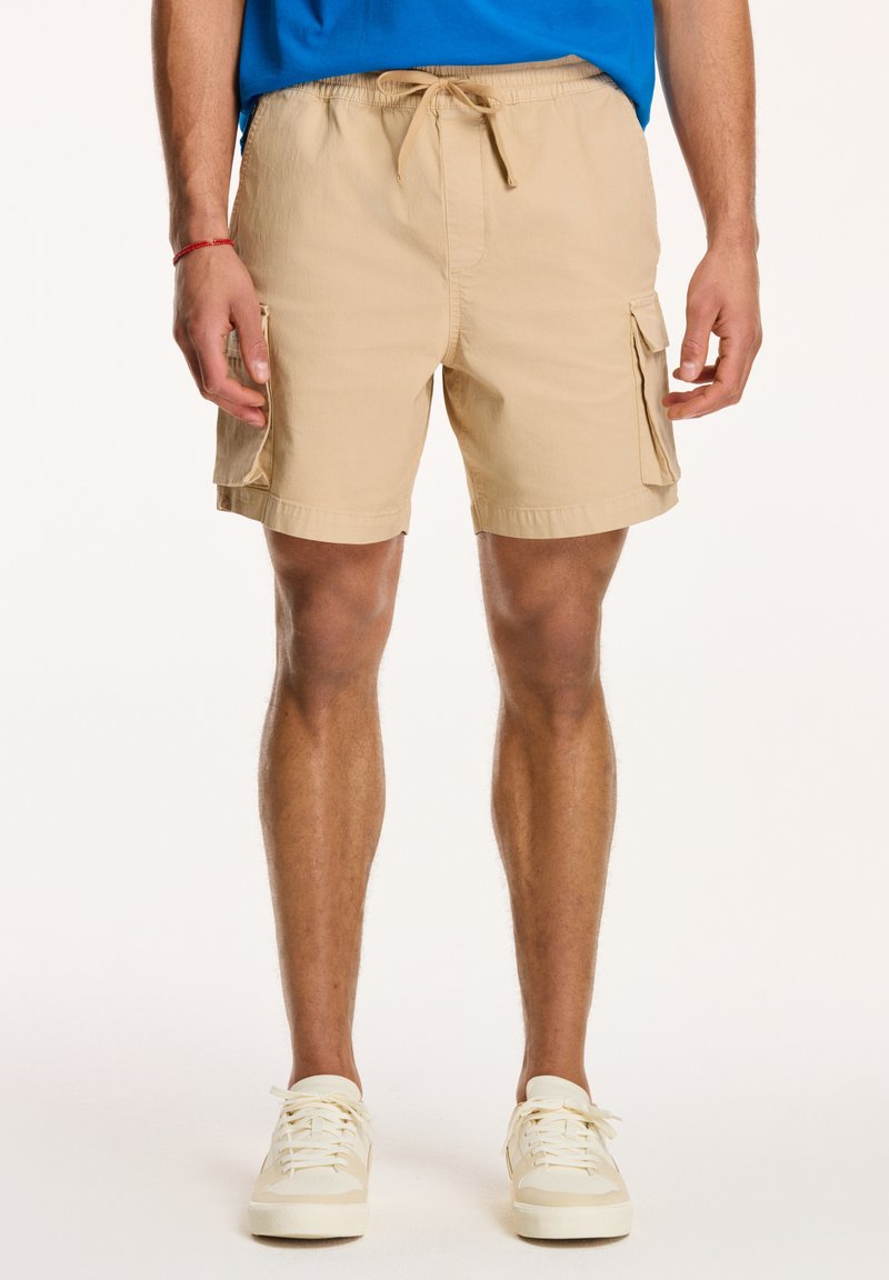 Shorts cargo bej cu talie ajustabilă prin sfoară, buzunare laterale și un material neted, ușor. Purtați cu adidași albi.