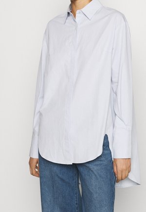 Osoba nosząca luźną białą, długą koszulę zapinaną na guziki z rozcięciami po bokach, w połączeniu z niebieskimi dżinsami na jednolitym tle.