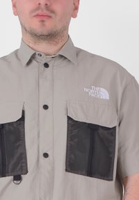 Maglietta grigia chiara a maniche corte con tasche in rete nere, apertura con bottoni e logo ricamato sul lato sinistro del petto. Tessuto liscio, design casual.