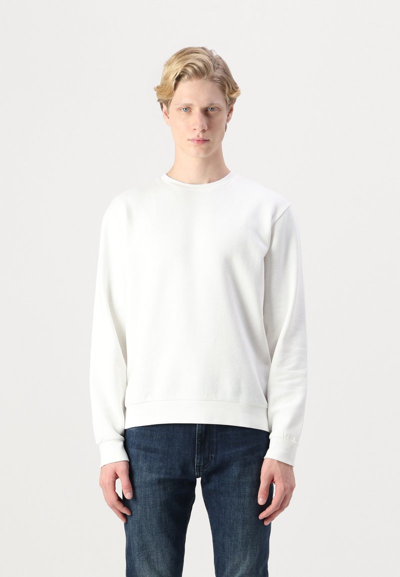 White crewneck sweatshirt made of a textured fabric, featuring long sleeves and a ribbed hem. Paired with dark blue denim jeans.