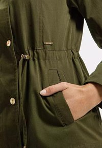 Oliwkowa kurtka z metalowymi detalami w odcieniu złota, elastycznym pasem i bocznymi kieszeniami. Wykonana z miękkiego, teksturowanego materiału o luźnym kroju.