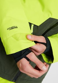 Personne portant une veste O'Neill jaune-vert et gris foncé ajustant le poignet Velcro sur une manche noire avec des trous pour les pouces.