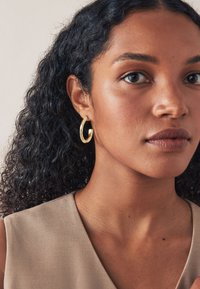 Gold hoop earring with a textured surface, worn on a person with curly hair and a neutral-colored sleeveless top, featuring subtle skin texture.