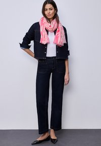 Femme portant une veste et un pantalon en denim foncé, une chemise blanche, une écharpe rose à motifs, et des ballerines noires pointues, debout contre un mur gris clair.
