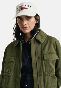 Olivgrüne Jacke mit zwei Brusttaschen, ausgestattet mit einem Reißverschluss und Druckknöpfen. Hellfarbige Mütze mit "GANT USA" in Rot und Marineblau gestickt.