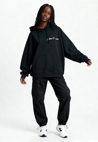 Sweat à capuche noir surdimensionné avec poche avant et texte blanc "je m'en fiche" ; associé à un pantalon cargo noir et des baskets blanches avec des accents noirs.