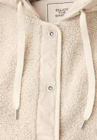 Gros plan sur une veste en polaire couleur crème avec des boutons-pression en métal et des cordons de serrage crème sur la capuche, montrant la texture du tissu et le texte de l'étiquette.