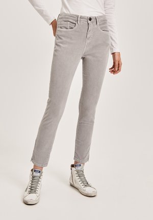 Personne portant un pantalon slim en velours côtelé gris clair et des baskets montantes blanches avec des lacets gris, debout devant un fond uni.