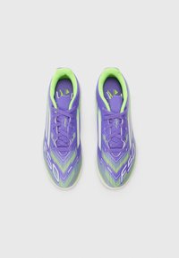 Botas de fútbol moradas con un diseño estructurado, que presentan detalles verdes, una superficie texturizada y un patrón geométrico. Lazos y logotipo visibles.